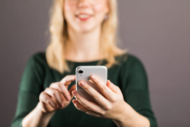 Junge Frau - Kopf oben im Anschnitt - hält vor sich ein Smartphone in der linken Hand, mit dem Zeigefinger der rechten Hand berührt sie fast das Display.