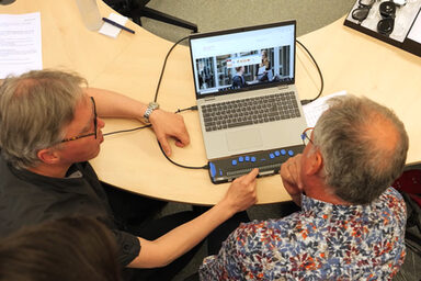 Two people are sitting at a table. And they are looking at a laptop. One person shows the other a device that blind people can use to capture the contents of the screen.