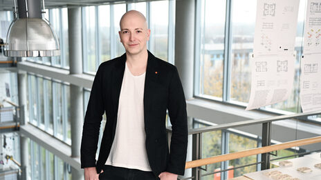 Ein Mensch im schwarzen Sakko steht in einem Gebäude an einem Geländer vor einer Fensterfront.