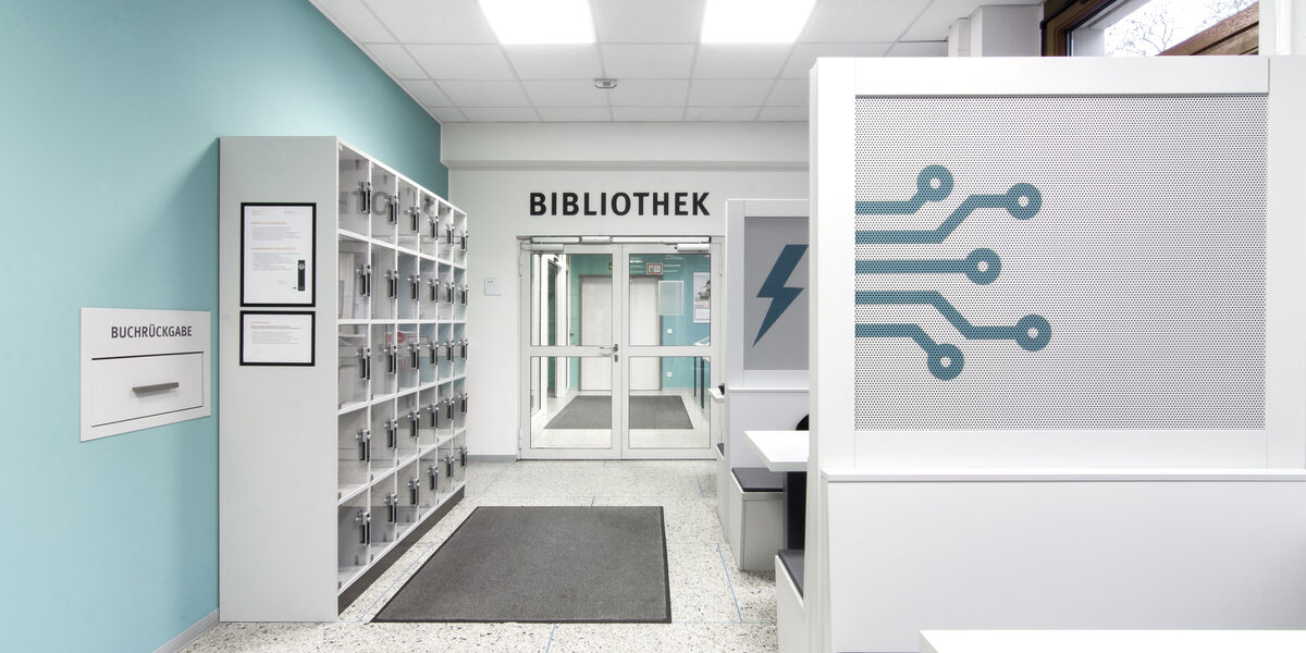 Foto des Flurs vor dem Eingang zur Bibliothek. Dort befinden sich Schließfächer, eine Rückgabebox und Gruppenarbeitsplätze__Photo of the hallway in front of the library entrance. It features lockers, a book return box and desks for group study