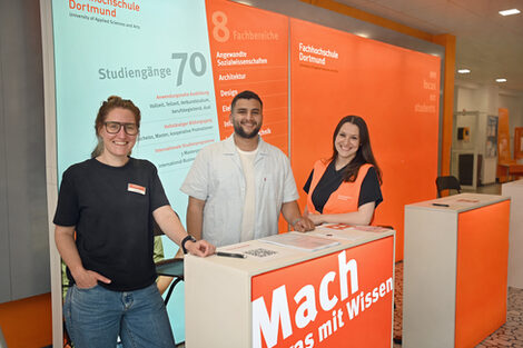 Three people are standing in front of an orange wall that reads "Do something with knowledge". They are smiling.