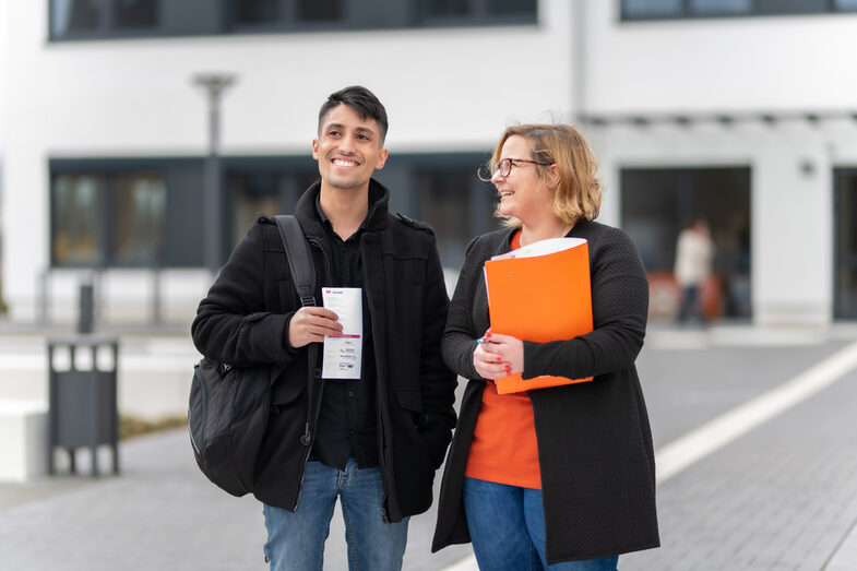 Foto von einer Mitarbeiterin und einem Schüler, die vor einem Campusgebäude stehen. Sie hält eine orangene Mappe unterm Arm, er hat einen Flyer in der Hand. Beide lachen.