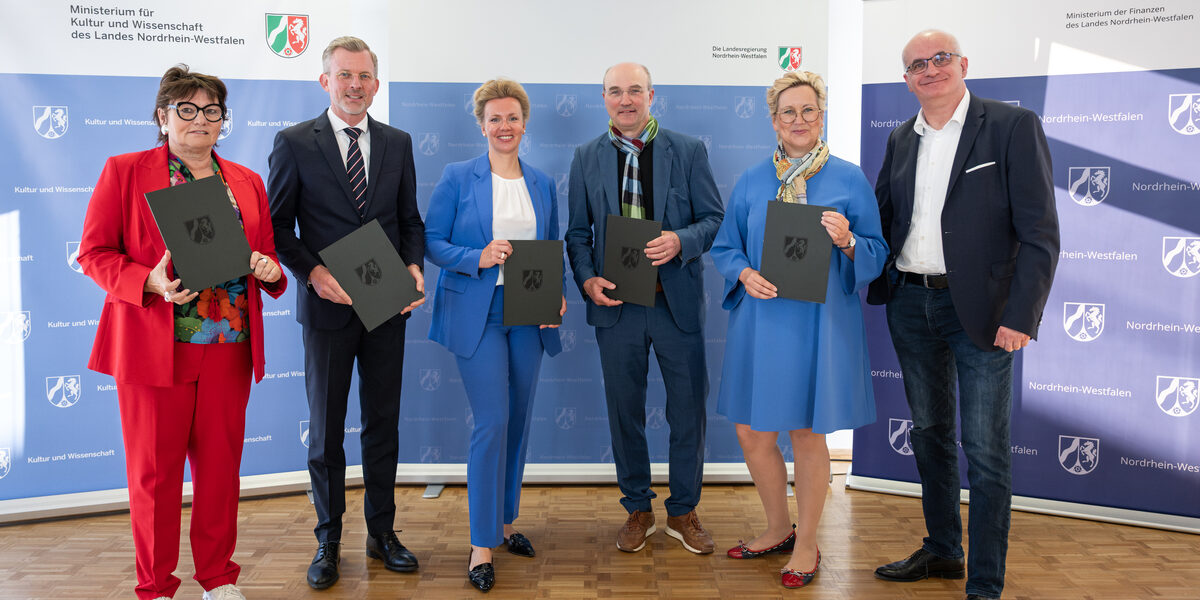 Sechs Personen stehen nebeneinander, fünf von ihnen halten eine Mappe vor sich.