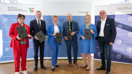 Sechs Personen stehen nebeneinander, fünf von ihnen halten eine Mappe vor sich.