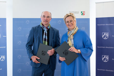 Zwei Personen stehen nebeneinander und halten jeweils eine Mappe vor sich.