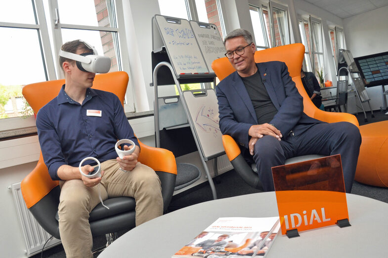 Two people are sitting on two armchairs next to each other. The UAS employee on the left has put on VR glasses and is holding two control elements, while the Lord Mayor looks on.