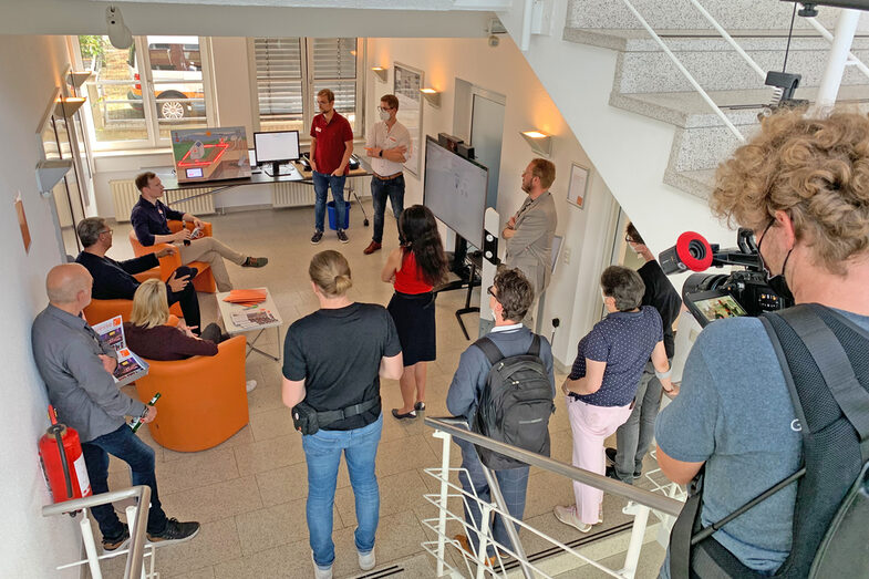 Several people sit and stand on a floor in the hallway to watch a presentation on a screen. A cameraman is documenting what is happening.