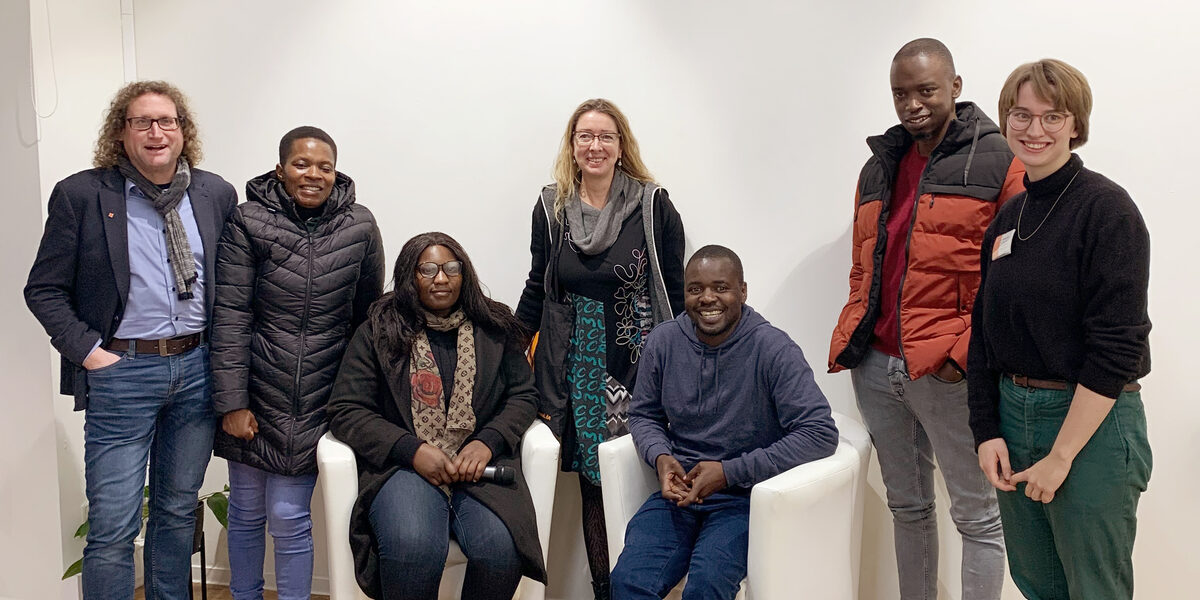 Seven people, some standing, some sitting, in front of a white wall look into the camera.