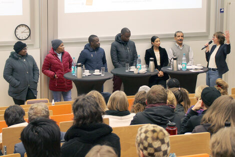 Sieben Personen stehen in einem Hörsaal vor Publikum, die Person ganz rechts spricht in ein Mikrofon und gestikuliert.