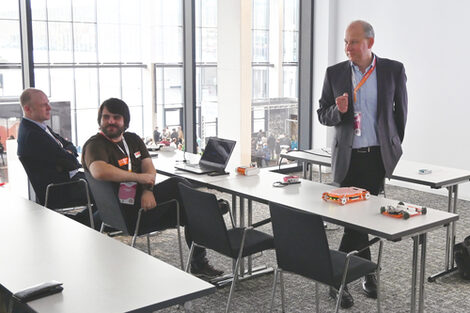 Two people are sitting at a row of tables, one person is standing in front of them. There are various objects on the tables, including a laptop and mobile models.