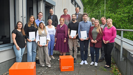 Eine Gruppe von Menschen steht um orangefarbene Würfel. Einige von ihnen halten Urkunden.