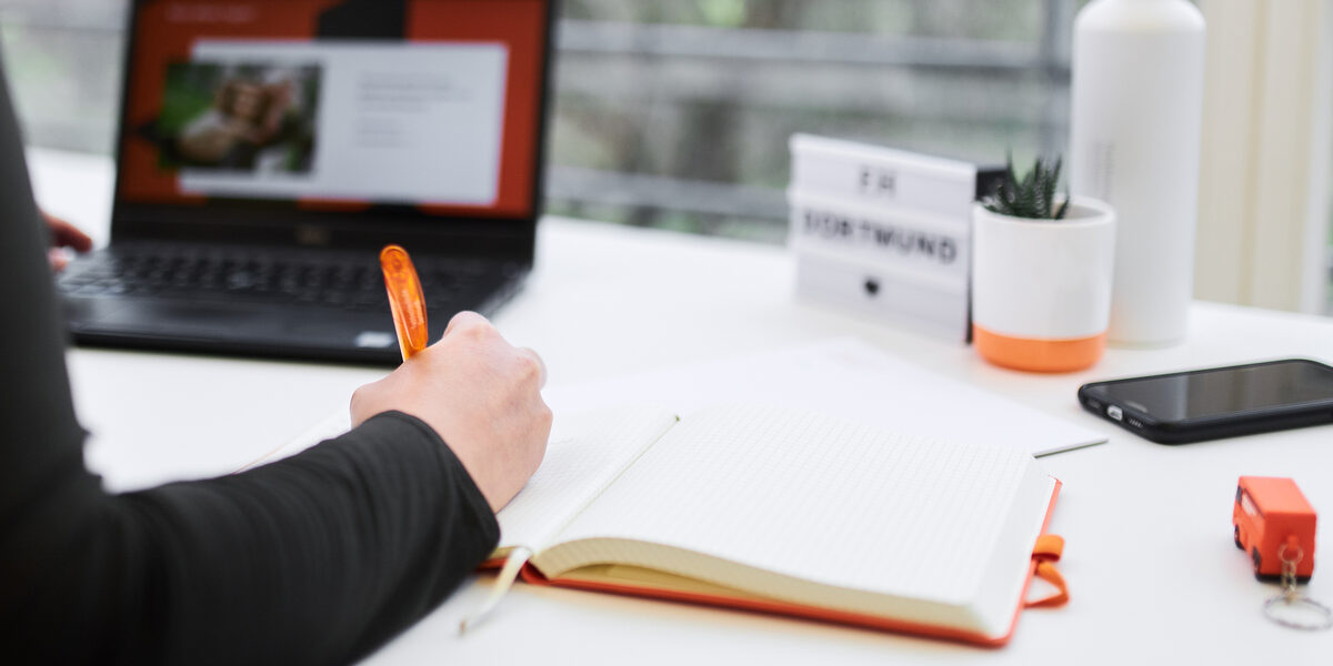 Photo of a workplace with a laptop. In the foreground you can see the arm of a person writing something in a notebook.