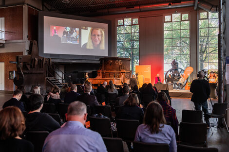 The audience sits in front of a stage on which three people can be seen. To the left of the stage is a large screen showing a close-up of the stage as well as a person who has been switched on hyprid.