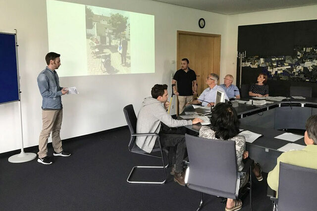 Study on quality of life. People sit and stand around a group of tables and look ahead at a presentation.