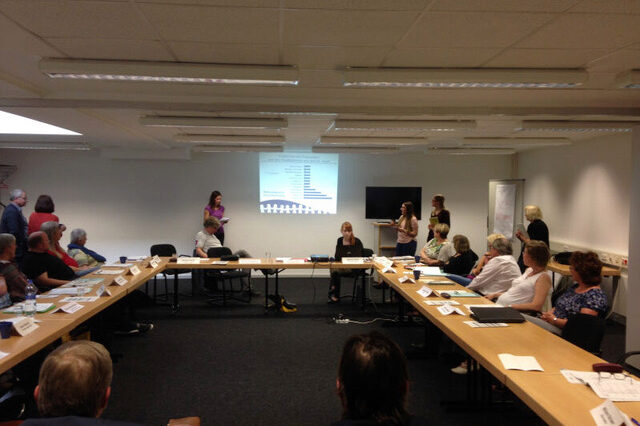 Conference on ageing and care. People sit and stand around a group of tables and look ahead at a presentation.