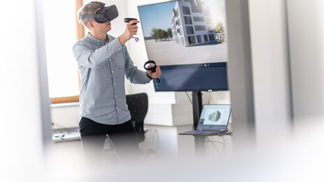 Photo of a man with VR glasses on and controller in his hand. On the screen in the background, you can see a building in the perspective that the man sees through the VR glasses.