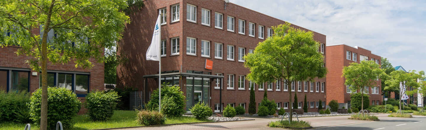 Photo of the location of the Fachhochschule Dortmund in Otto-Hahn-Straße from the outside. It is a red brick building. In the foreground are a road and some trees.