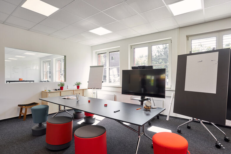 Raumaufnahme mit einem Konferenztisch, Flipchart, digitalem Board und Notizwand.