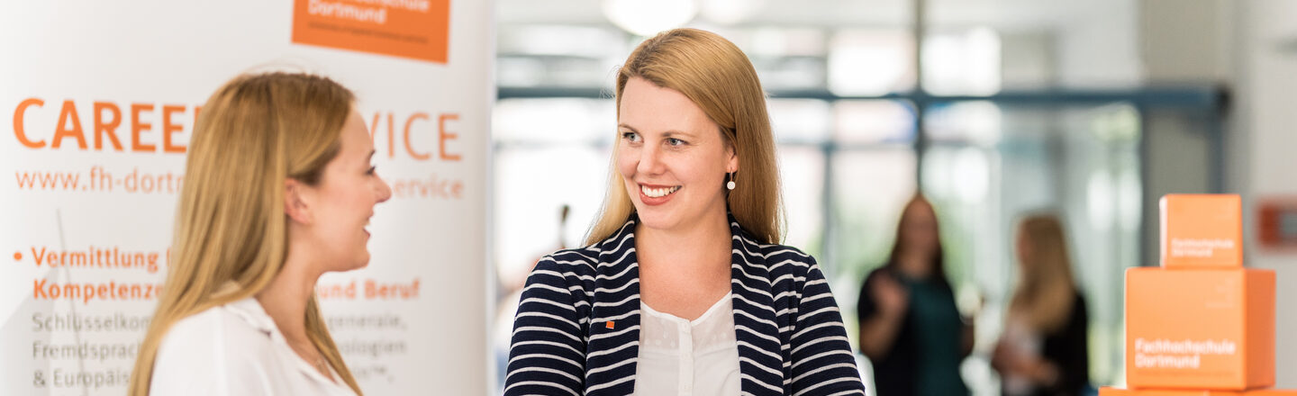 Photo of an employee of the Career Services of the Fachhochschule Dortmund standing at a high table talking to a young woman. __ <br>Employee of the Career Services of the University of Applied Sciences Dortmund stands at a high table talking to a young woman.