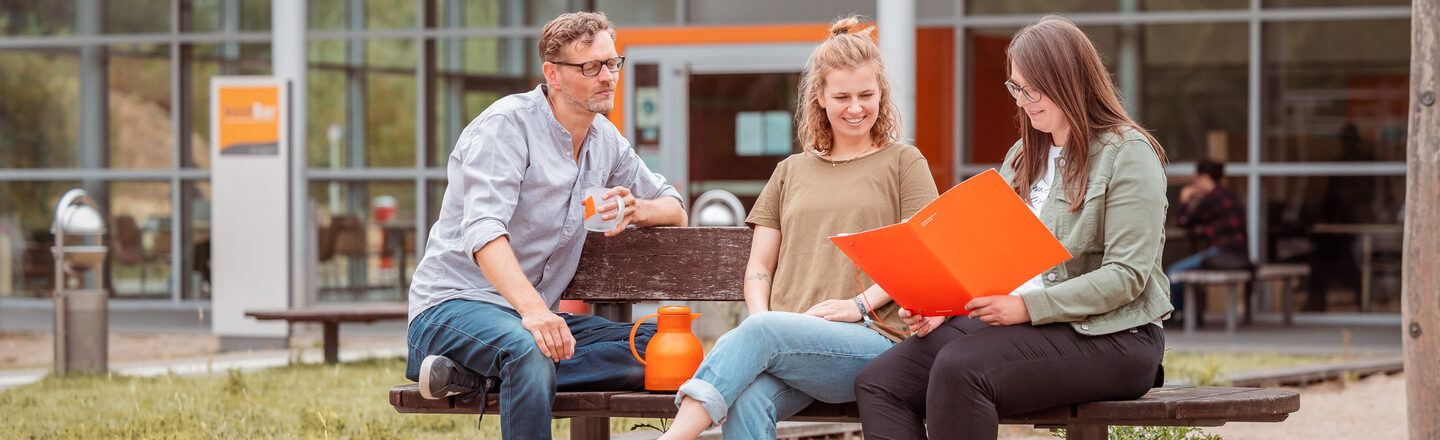 Foto von 3 Personen, die draußen vor der kostBar auf einer Holzbank sitzen und sich besprechen.