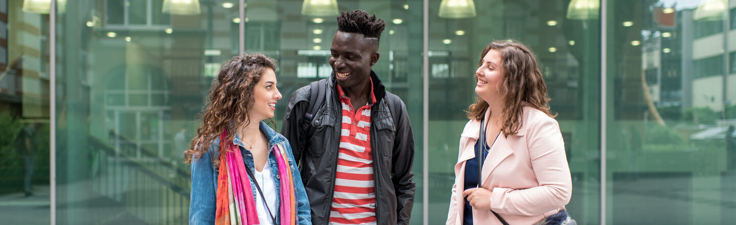 Foto von zwei Studentinnen und einem Studenten, die nebeneinander vor dem Mensagebäude stehen. Sie lachen einander an.__Two female and one male students stand side by side in front of the canteen building, they laugh at each other.