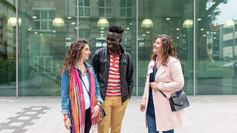 Photo of two female and one male students standing side by side in front of the canteen building, they laugh at each other. They laugh at each other.__Two female and one male students stand side by side in front of the canteen building, they laugh at each other.