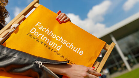 Foto von einer Studentin - stark im Anschnitt. Sie trägt eine orangefarbene Klappliege mit FH-Logo. __ Female student - strong in the cut - wears an orange folding chair with the FH logo.