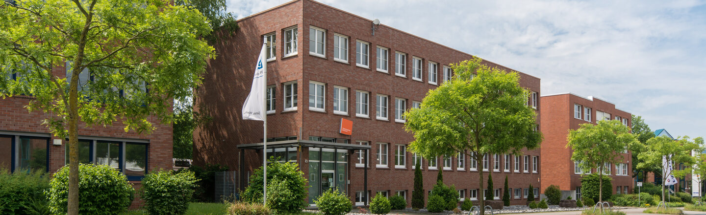 Foto des Standorts der Fachhochschule Dortmund in der Otto-Hahn-Straße von außen. Es ist ein rot verklinkertes Gebäude. Im Vordergrund sind eine Straße und einige Bäume.