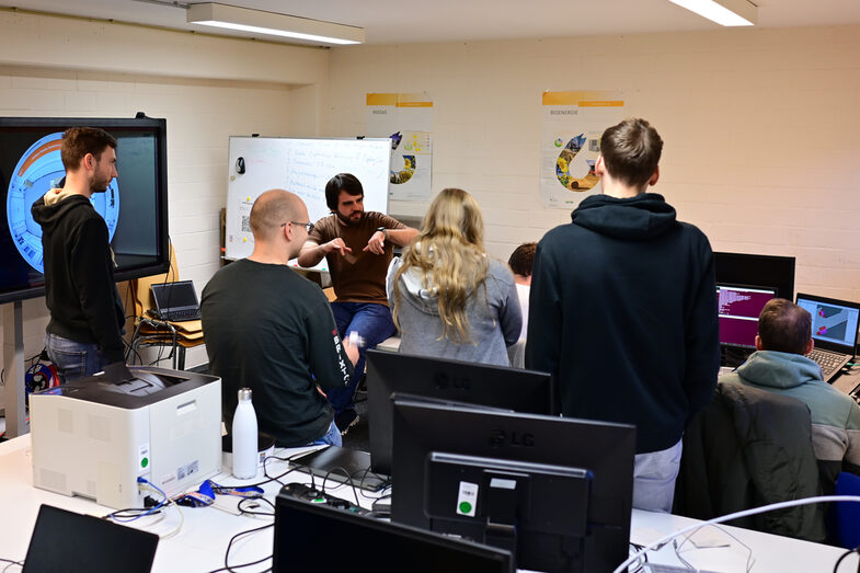 Studierende sind im Studierendenarbeitsraum des IDiALs versammelt. Die Blicke sind auf Alexander Miller gerichtet, der stark gestikulierend die Kinematik der EduRobs erklärt.