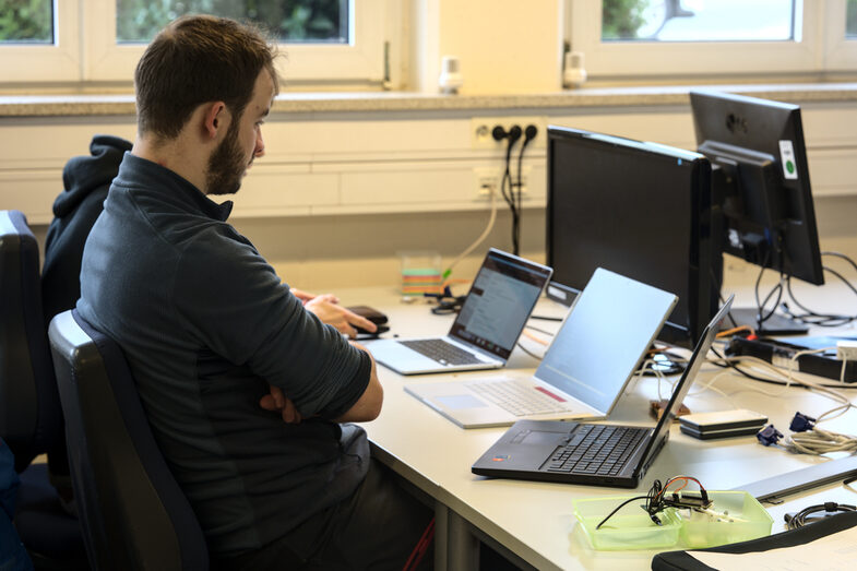 Two block week participants sit together at a table in front of a notebook and discuss the interface to the gripper. The gripper is lying next to the notebook in a lunch box on the table.