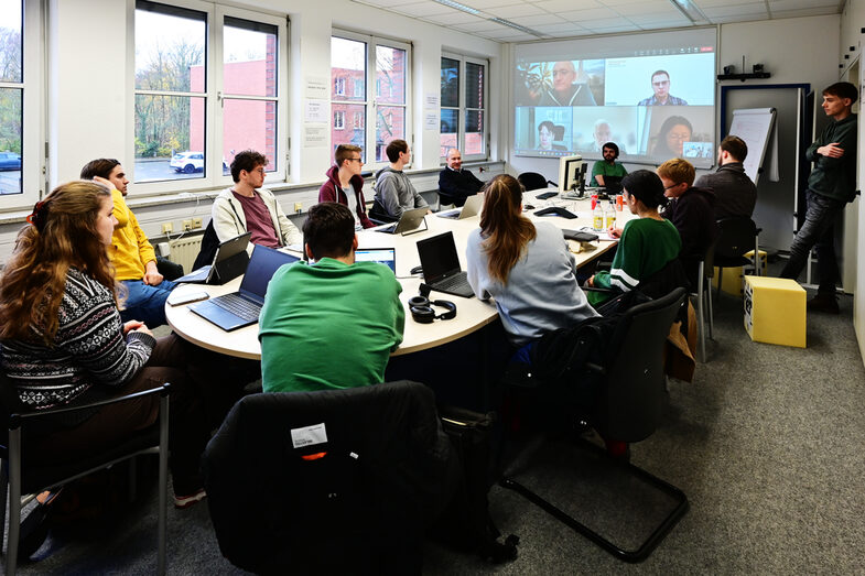 Studierende sitzen im Besprechungsraum des IDiALs um den großen Tisch und diskutieren mit den per Video-Konferenz zugeschalteten Professoren den Ablauf des letzten Tages der Blockwoche.