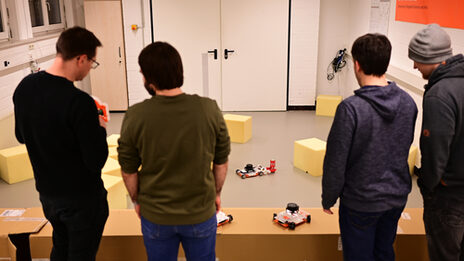 Four people (seen from behind) stand in front of the scenario set up in the robot lab. An EduRob is gripping a drinks can using the gripper developed during the block week. Two people are discussing an EduRob held by the person on the left.