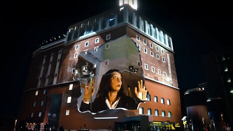 Projection onto the façade of the Dortmunder U: A woman presses her face against an invisible pane of glass in surprise.