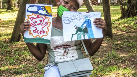 A girl holds up two pictures painted by children. The pictures focus on the coronavirus pandemic and the topic of vaccination.