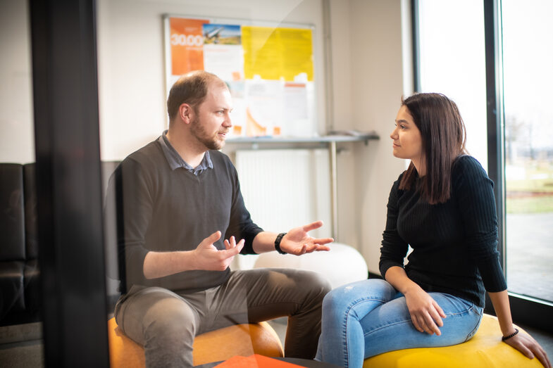 Foto von einem TalentScout und einer Schülerin, die auf Sitzsäcken sitzen und sich unterhalten. Vor ihnen liegen auf einem Beistelltisch einige orangefarbene Mappen.