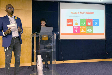 Two people are standing at and next to a lectern, the person on the left is speaking. A projector image can be seen in the background.