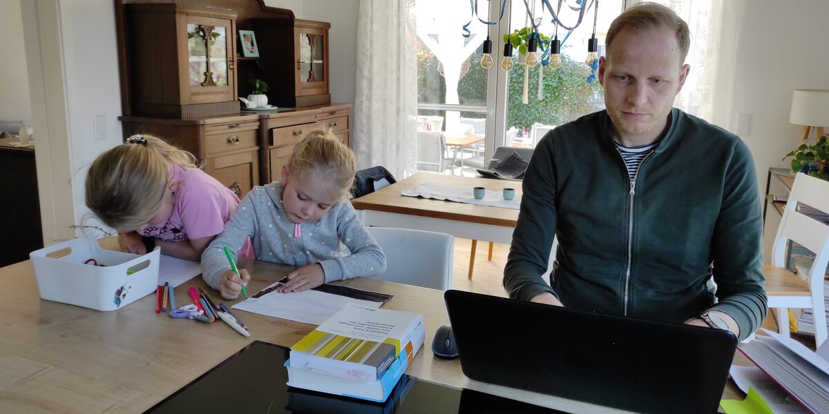 Scholarship holder Jens Terbrack is sitting at the table working on his laptop. Next to him, his two daughters are painting pictures.