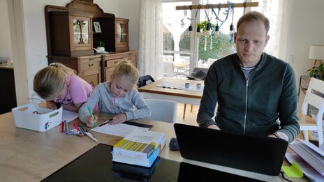 Scholarship holder Jens Terbrack is sitting at the table working on his laptop. Next to him, his two daughters are painting pictures.