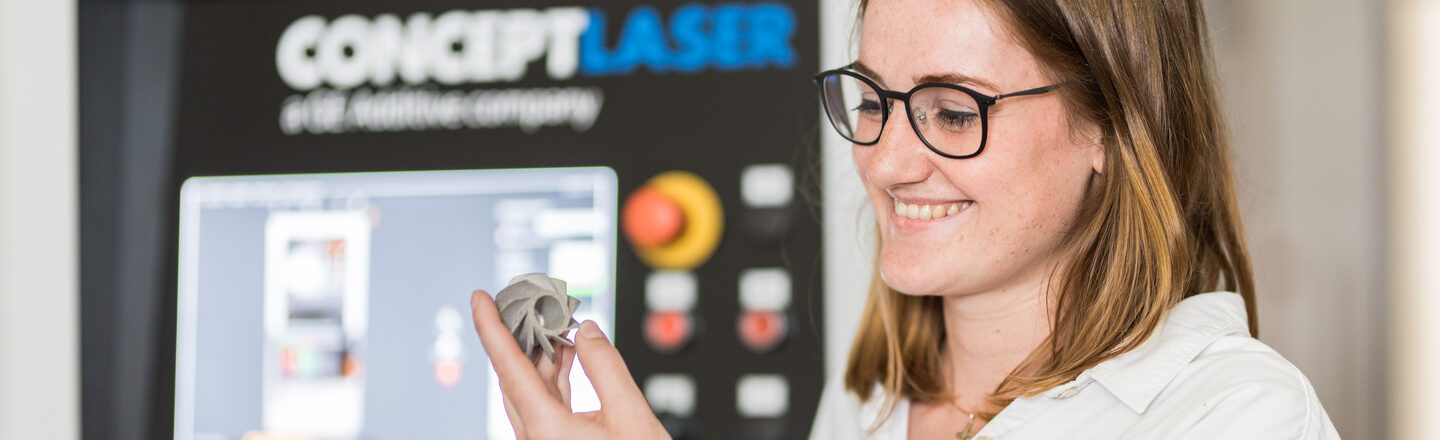 Photo of a woman looking at a 3D printed metal model in her hand, the printer can be seen out of focus in the background.