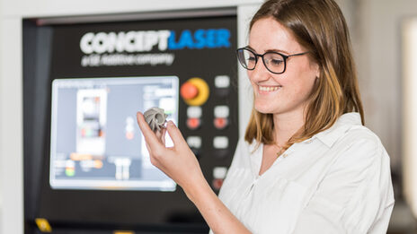 Photo of a woman looking at a 3D printed metal model in her hand, the printer can be seen out of focus in the background.