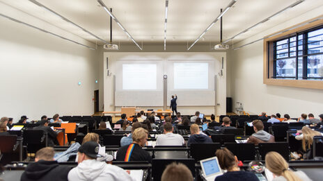 Von hinten Blick in einen vollen Hörsaal, vorne schreibt ein Lehrender etwas an die Tafel.