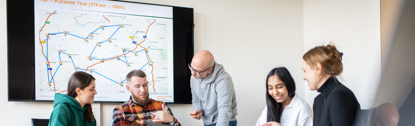 Photo of several students sitting at a table. A lecturer is standing in the middle of them, leaning down and explaining something.