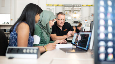 Foto von zwei Studentinnen und einem Professor im Labor für Energieautomation und Netzführung. Sie sitzen nebeneinander und schauen gemeinsam auf den Bildschirm eines Laptops.