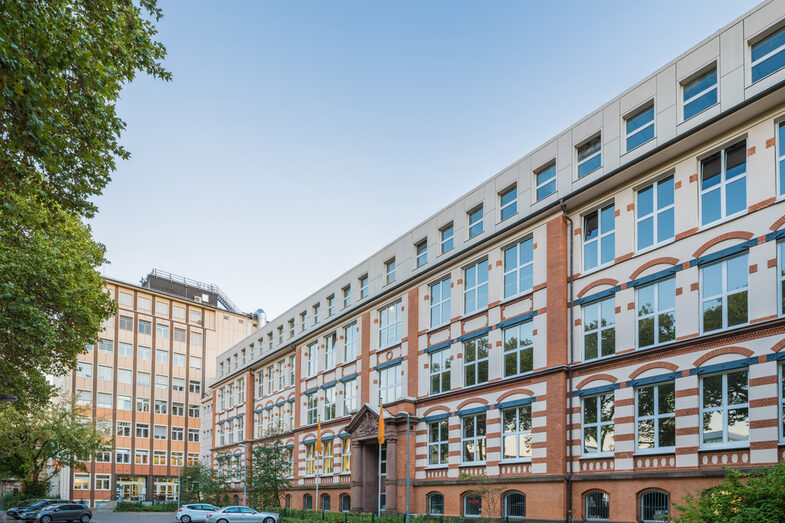 Foto des Altbaus und des Hochhauses an der Sonnenstraße mit Parkplatz davor. __ <br>View of the old building and the skyscraper on Sonnenstrasse across the parking lot.