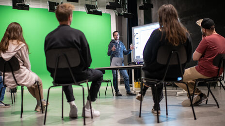 Photo of a man standing in front of a circle of students, 4 students can be recognized. The students are looking at the man.