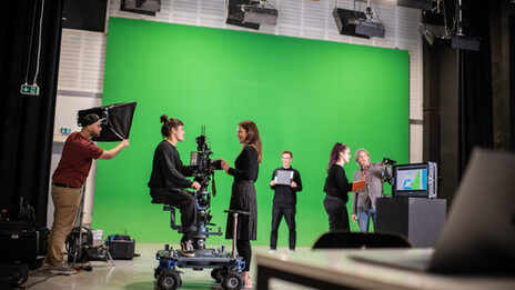 You can see a film studio with a green screen in the background. On the far left, a man is holding a light screen, to the right, two women are talking to a camera. Next to them is a man in front of a green screen as a model. On the right, a man and a woman are at a screen on which the film footage can be seen__A film studio can be seen with a green screen in the background. On the far left a man is holding a light screen, and to the right two women are talking at a camera. Next to it is a man in front of the green screen as a model. In the right part are a man and a woman at a screen showing the film footage