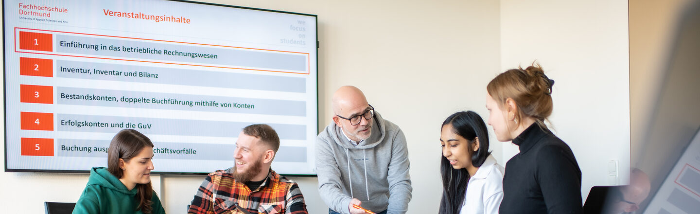 Foto von mehreren Studierenden, die an einem Tisch sitzen. In der Mitte von ihnen steht ein Dozent, der sich zu ihnen runter beugt und etwas erklärt.