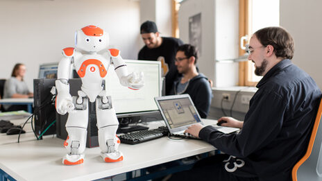 Photo of the NAO robot Eva standing on the table in front, arms raised. Next to it, a student sits at the laptop to give Eva commands.