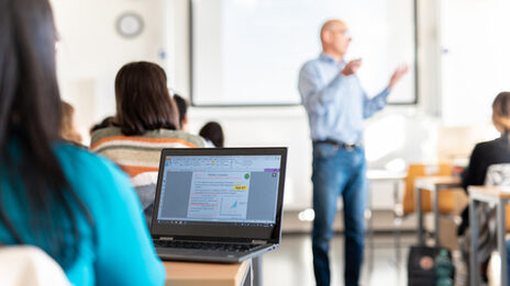 View of a student's laptop in a row of seats. Blurred in front of her, a lecturer explaining something.