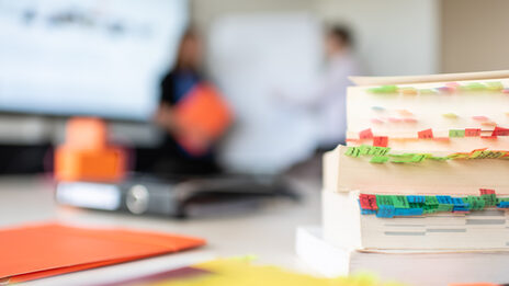 A pile of books with many different colored notes on the table as well as other documents. In the background, people in conversation are indistinctly recognizable.